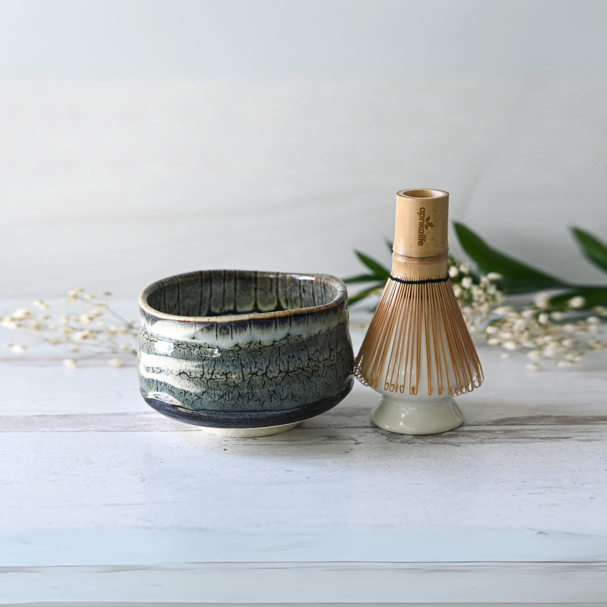 A Grey Tenmoku Ceramic Matcha Bowl and a bamboo matcha whisk rest on a light surface, with lush greenery in the background, creating a serene and inviting scene.