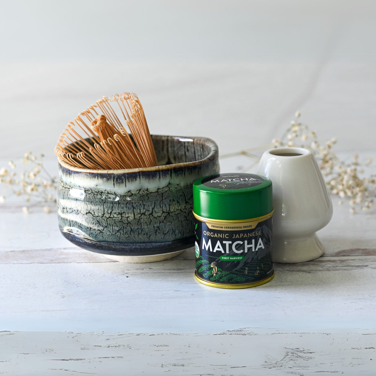 On a light wooden surface sits a Grey Tenmoku Ceramic Matcha Bowl, alongside a bamboo matcha whisk and a tin of organic Japanese matcha.