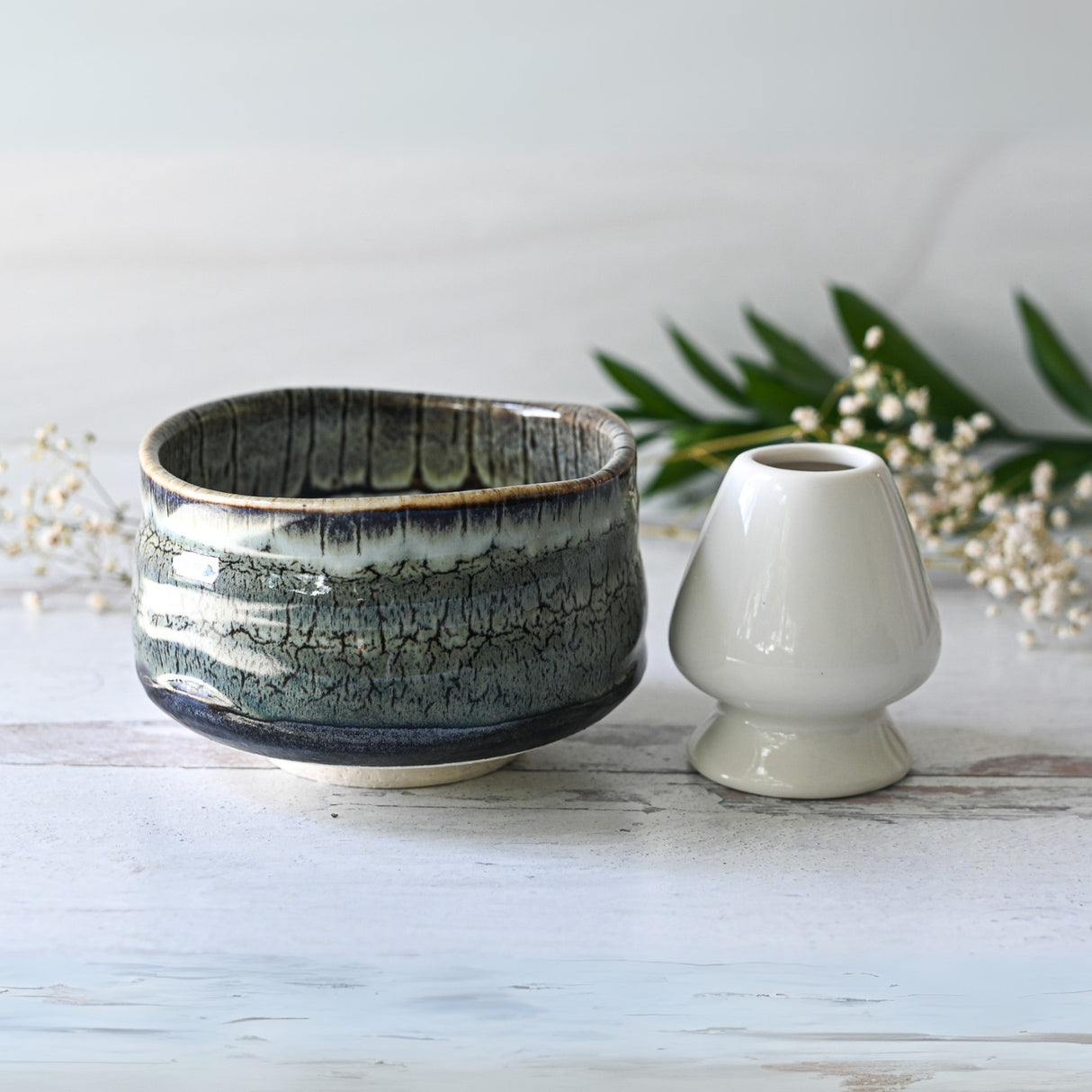 The Grey Tenmoku Ceramic Matcha Bowl with rustic glaze sits beside a small white vase on a light surface, foliage in the background creating a serene setting.