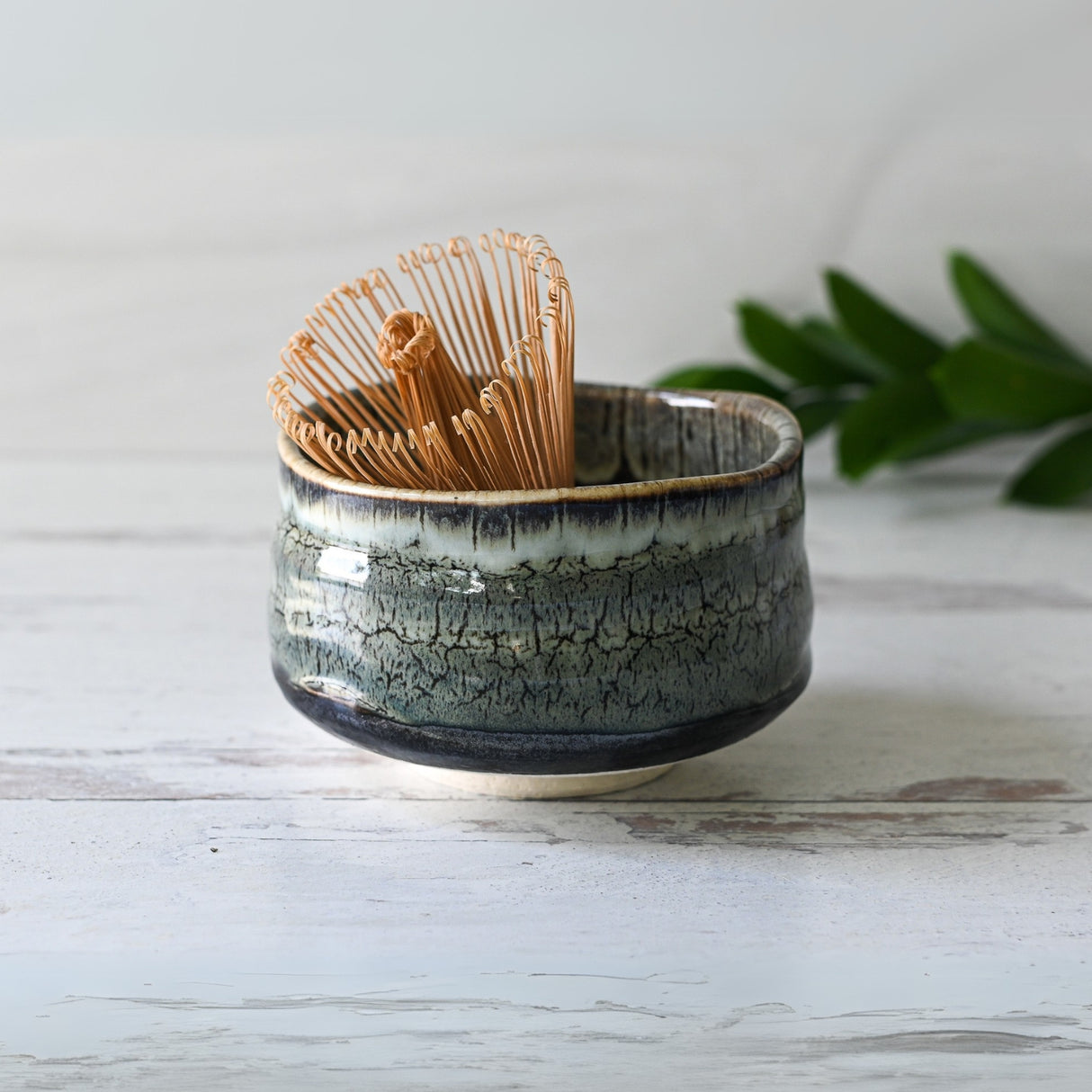 Grey Tenmoku Ceramic Matcha Bowl with Bamboo Matcha Whisk rests on a white wooden surface, framed by green leaves.