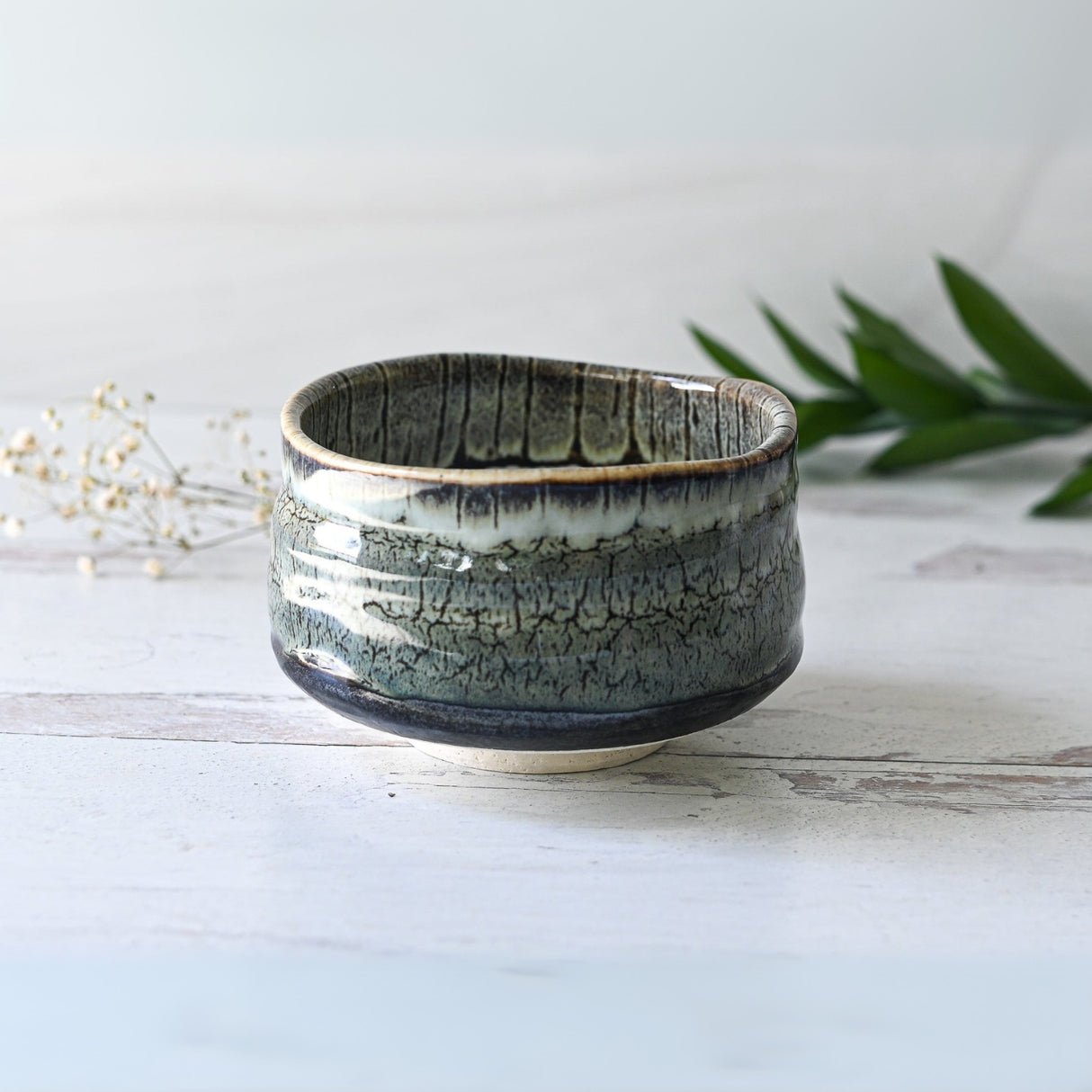 The Grey Tenmoku Ceramic Matcha Bowl, featuring a textured green glaze, sits on a light wooden surface adorned with delicate small flowers and leaves.