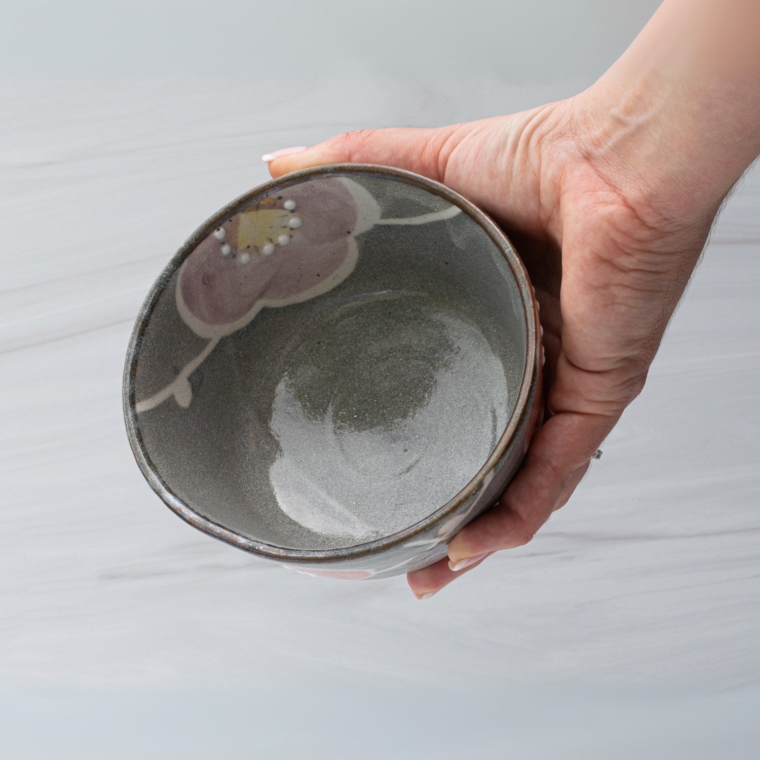 A hand holds the Gray with Flowers Ceramic Matcha Bowl, Bamboo Matcha Whisk and Whisk Holder Set—ideal tea accessories—against a light, textured background.