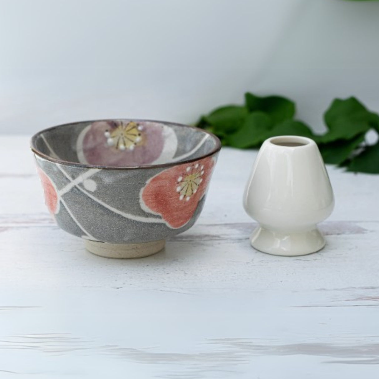 A Gray with Flowers Ceramic Matcha Bowl, Bamboo Matcha Whisk, and Whisk Holder Set are arranged on a light wooden surface with green leaves in the background.