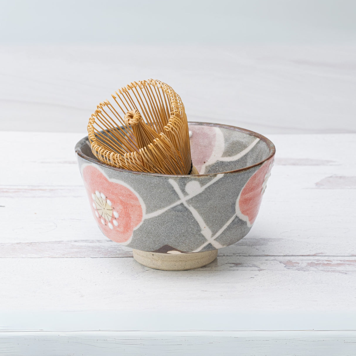 The Gray with Flowers Ceramic Matcha Bowl, Bamboo Matcha Whisk and Whisk Holder Set features a bamboo whisk inside a gray floral matcha bowl, beautifully arranged on a light wooden surface.