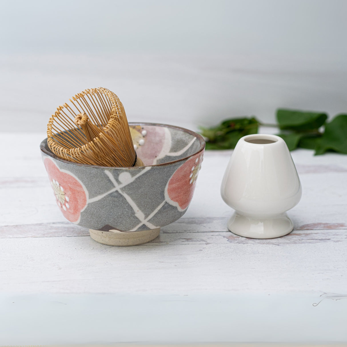 A handcrafted matcha set features a matcha whisk in a Gray with Flowers Ceramic Matcha Bowl, placed beside a small white holder on a light surface.