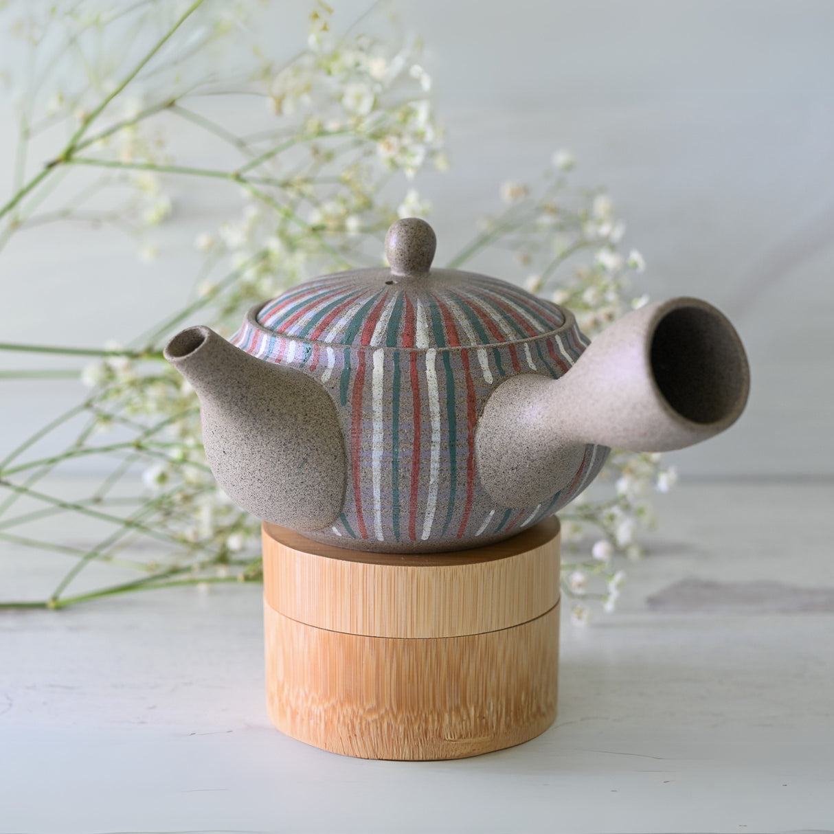 A Handcrafted Japanese Kyusu Teapot with Filters (310ml) featuring colorful stripes rests on a wooden stand, surrounded by delicate white flowers, highlighting the beauty of Japanese ceramics.