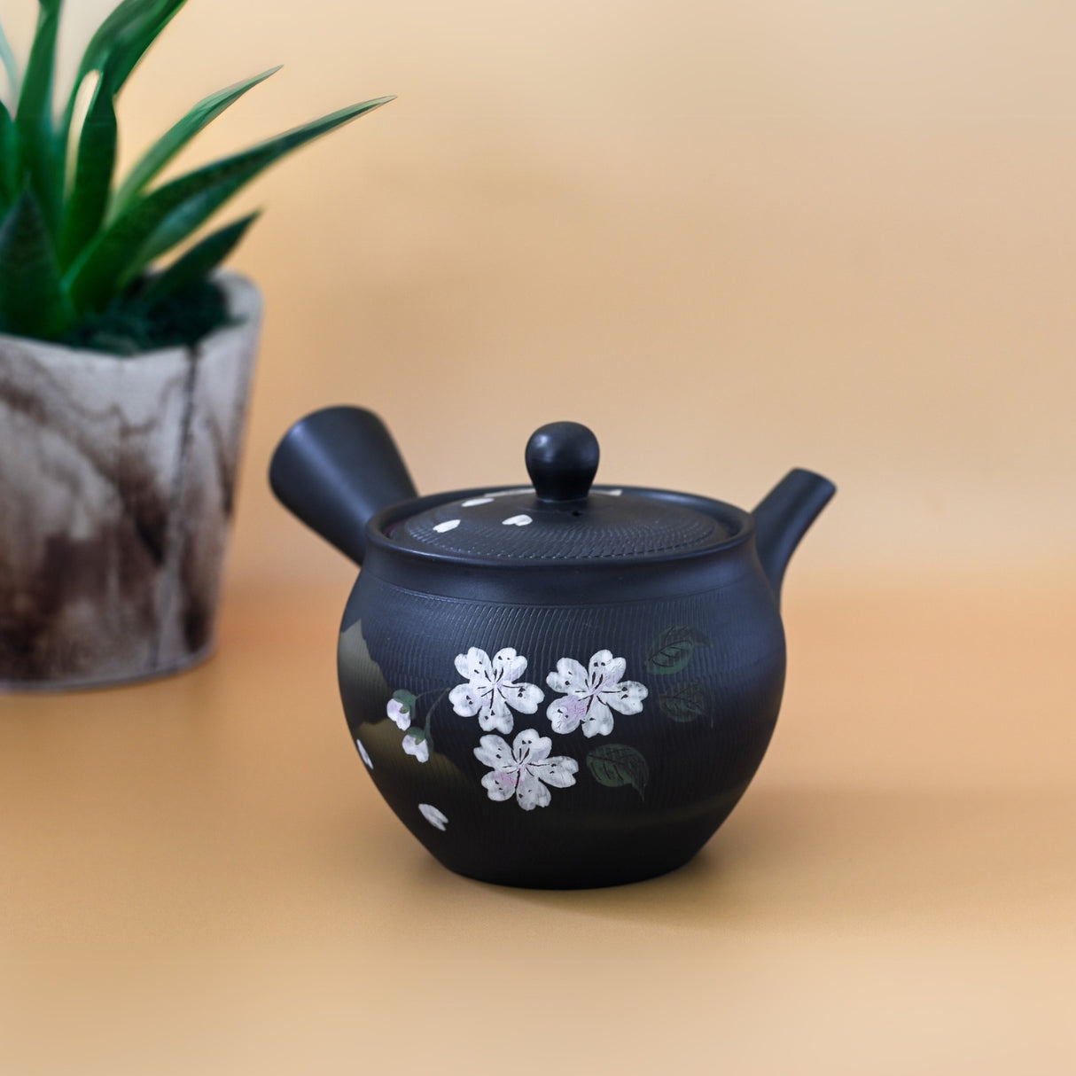 The Handcrafted Japanese Kyusu Teapot with Filters (350 ml), featuring exquisite black ceramic and floral designs, rests elegantly on a beige surface beside a small potted plant.