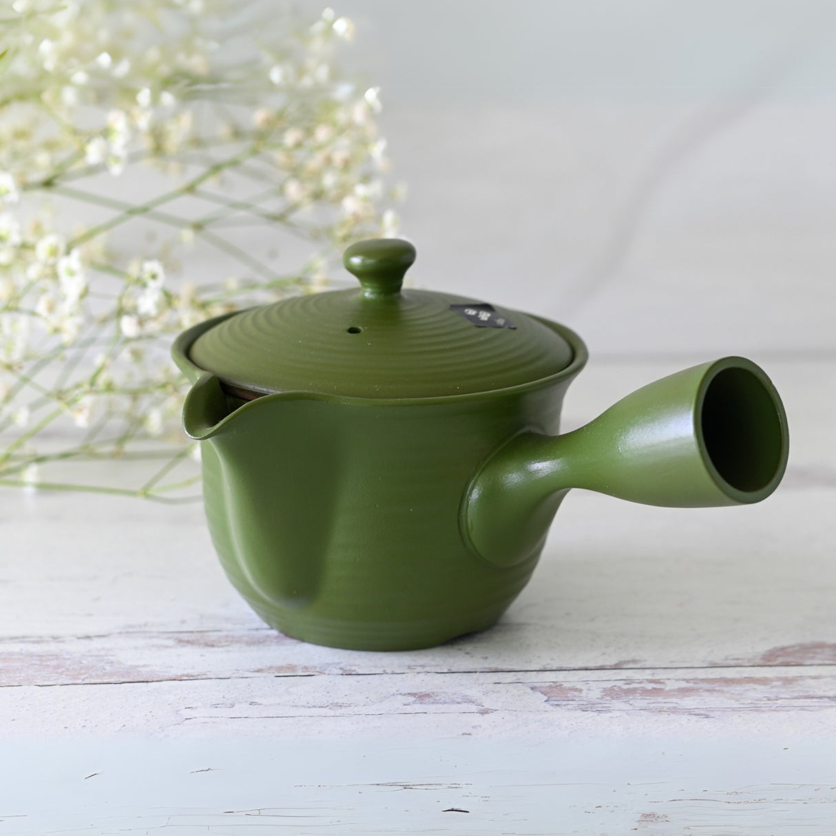 A beautiful green Handcrafted Japanese Kyusu Teapot with Filters (380ml) rests on a light wooden surface, surrounded by delicate white flowers. Inspired by Fujita Tokuta's mastery of Japanese ceramics, it features a ceramic strainer for the perfect brew.