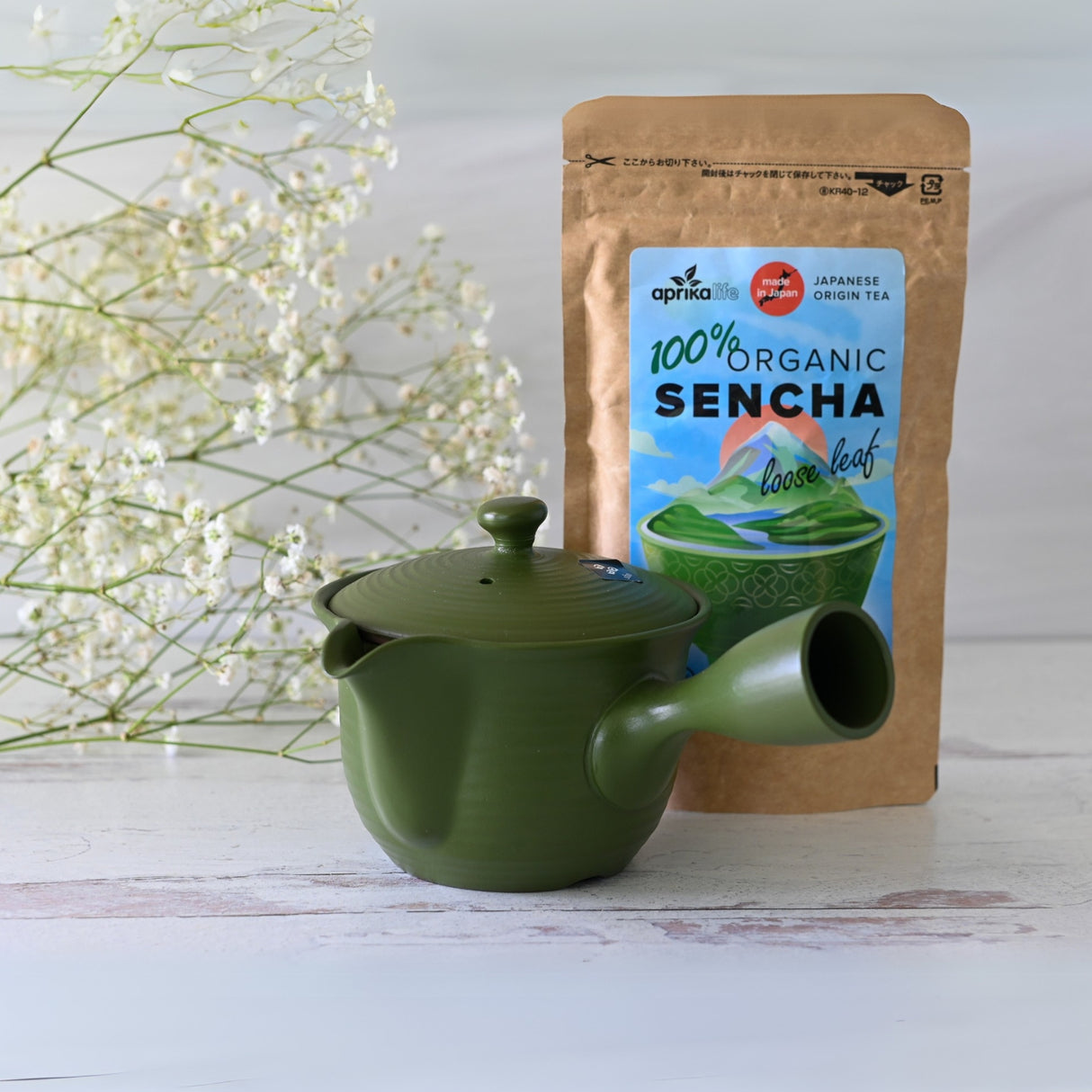 The Handcrafted Japanese Kyusu Teapot with Filters (380ml), in green, sits alongside a bag of organic sencha tea on a white surface, surrounded by delicate white flowers.