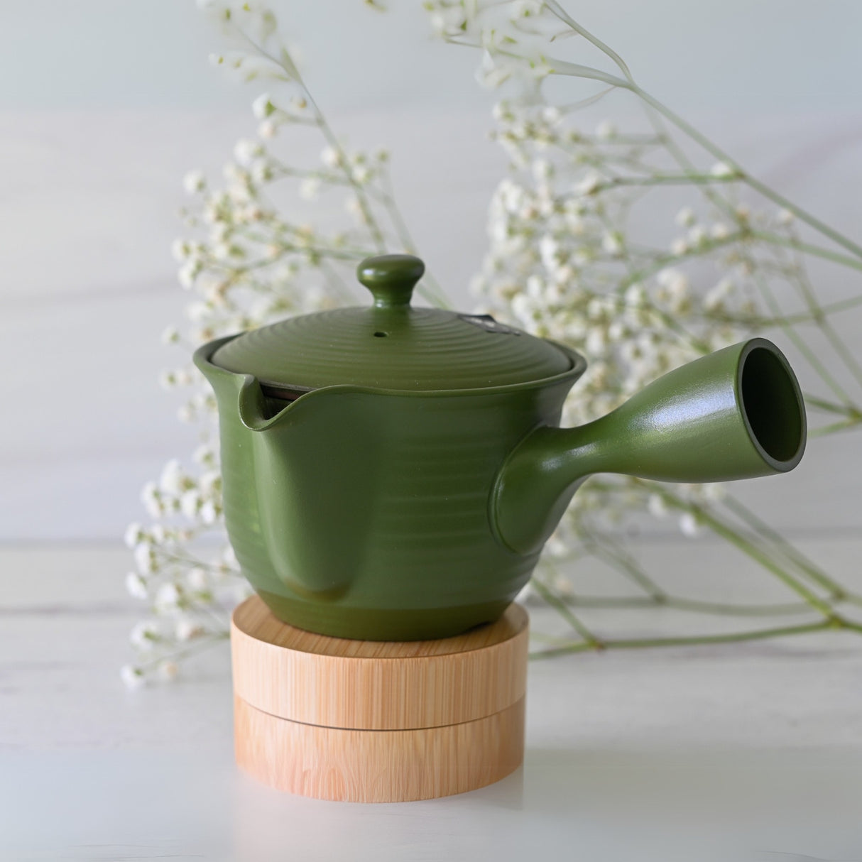 The Handcrafted Japanese Kyusu Teapot with Filters (380ml) in green, featuring a side handle, sits on a wooden stand surrounded by delicate white flowers against a light background.