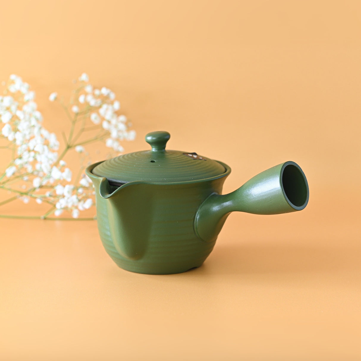 The Handcrafted Japanese Kyusu Teapot with Filters (380ml), inspired by Fujita Tokuta's ceramics, is shown in green with a side handle against a peach background, accompanied by small white flowers.