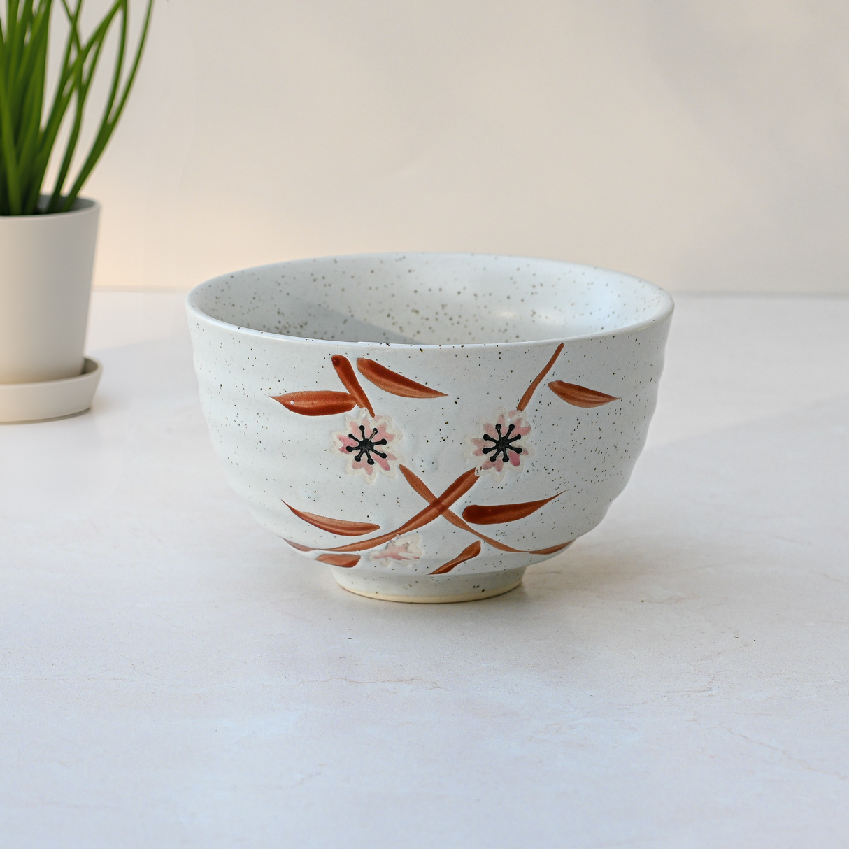 A Bamboo Matcha Bowl is displayed on a light surface, featuring a brown floral design, accompanied by a Bamboo Matcha Whisk from the Bamboo Matcha Whisk and Whisk Holder Set alongside a small potted plant.
