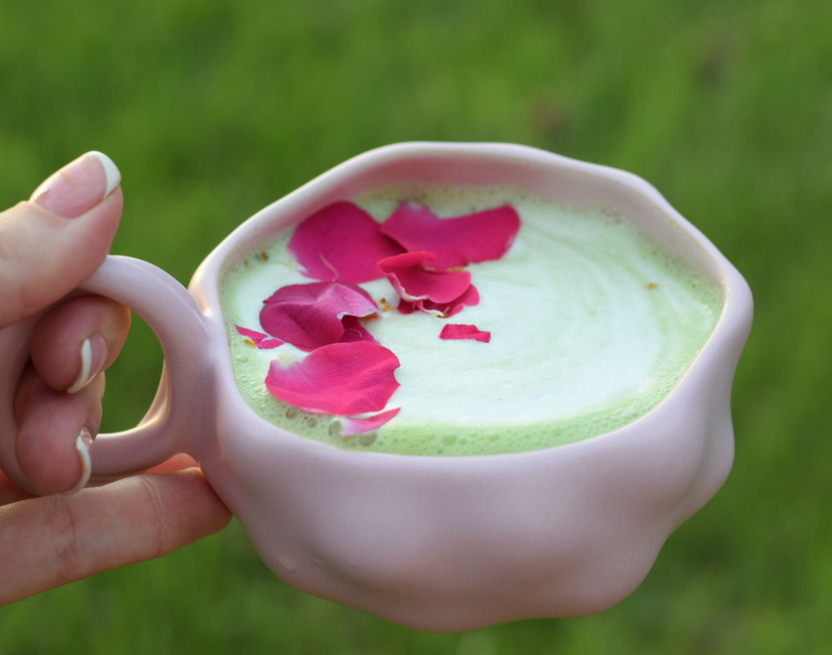 A hand holds a pink cup of green matcha latte made with Ceremonial Grade Matcha Latte Powder Mix with Natural Vanilla Flavor, topped with pink rose petals, set against a green background.