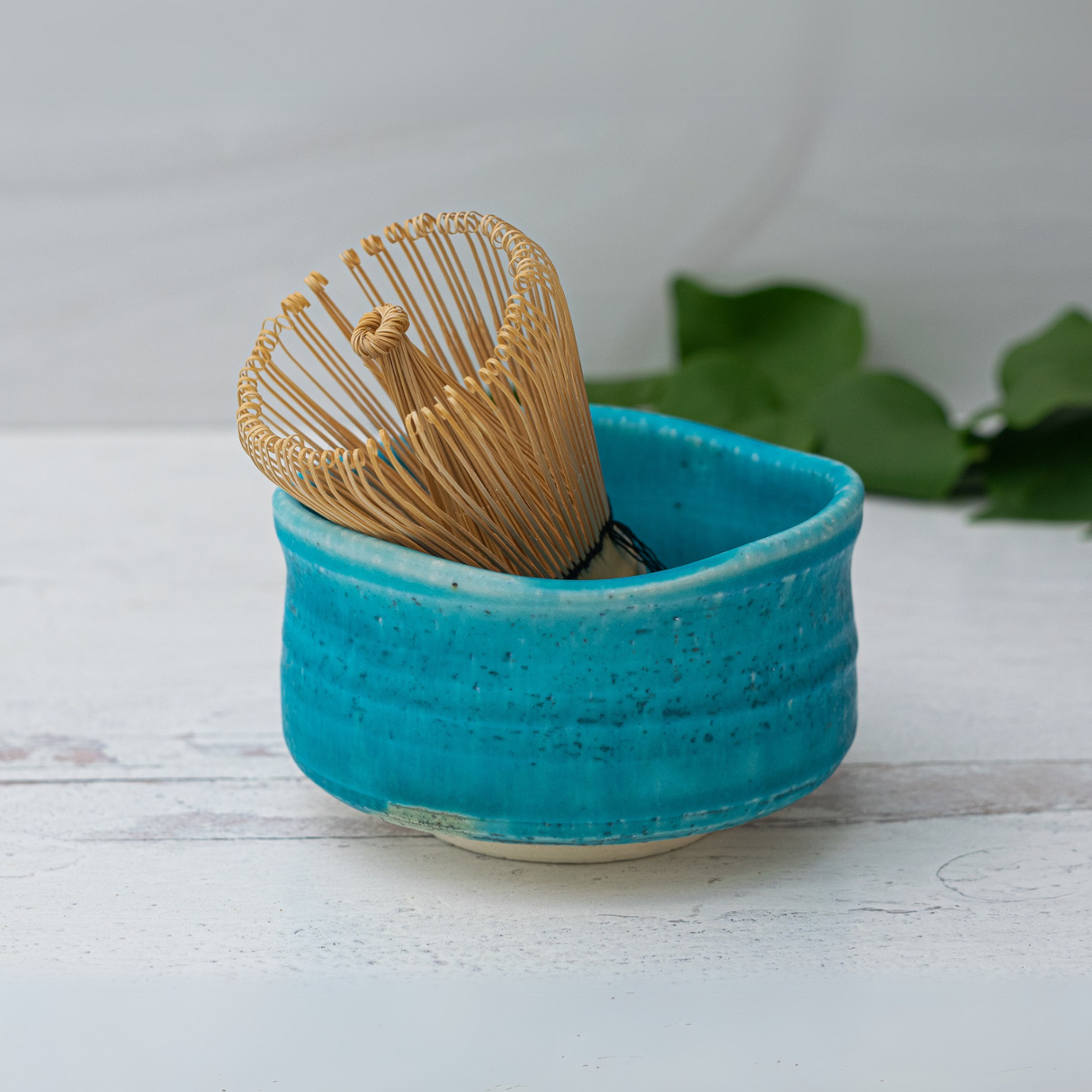 The Iguchi Blue Matcha Bowl, paired with a bamboo whisk, is elegantly displayed on a white wooden surface with green leaves in the background, highlighting the beauty of Japanese pottery.
