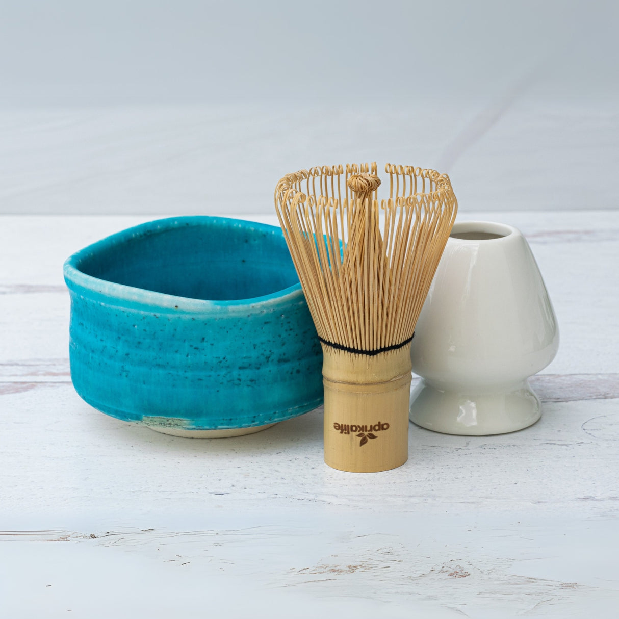 An Iguchi Blue Matcha Bowl, accompanied by a bamboo whisk and white holder, sits elegantly on a light wooden surface, highlighting the timeless beauty of Japanese pottery.