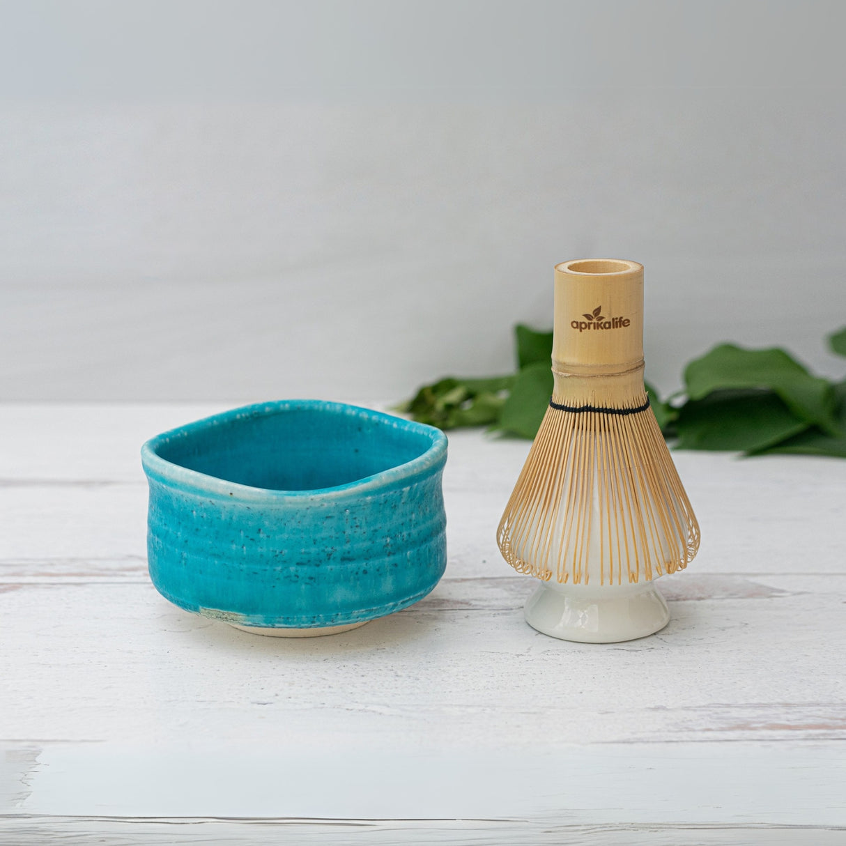An Iguchi Blue Ceramic Matcha Bowl and a bamboo whisk rest gracefully on a white surface, framed by green leaves in the background.