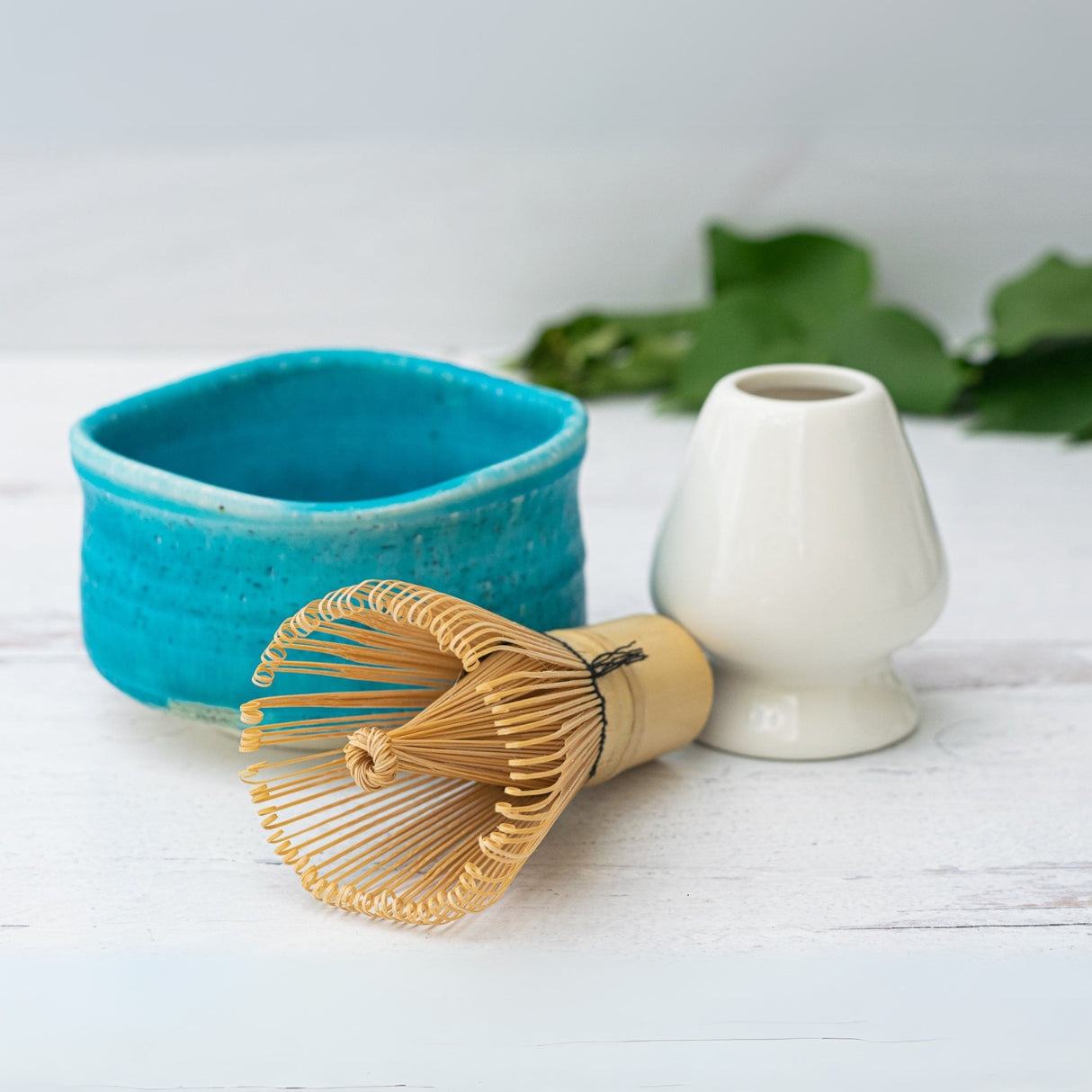 The Iguchi Blue Ceramic Matcha Bowl, accompanied by a bamboo whisk and white stand, rests elegantly on a light wooden surface, with green leaves in the background.