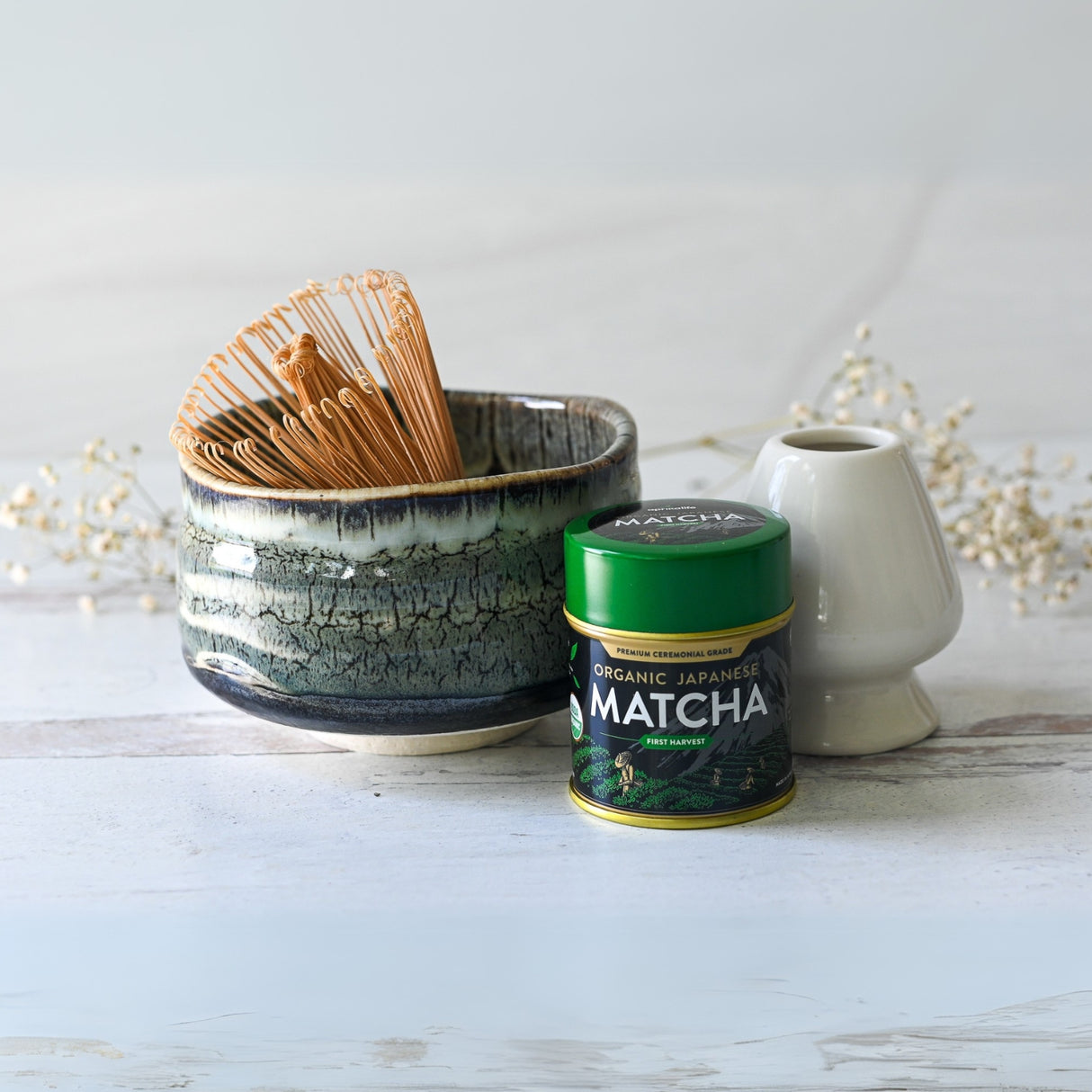 The Iguchi Tenmoku Glaze Matcha Bowl, Bamboo Whisk, and Organic Japanese Matcha Set sit on a light wood surface with a small white vase. Dried flowers are in the background, and the Tenmoku glaze offers an elegant touch inspired by traditional Minoyaki craftsmanship.