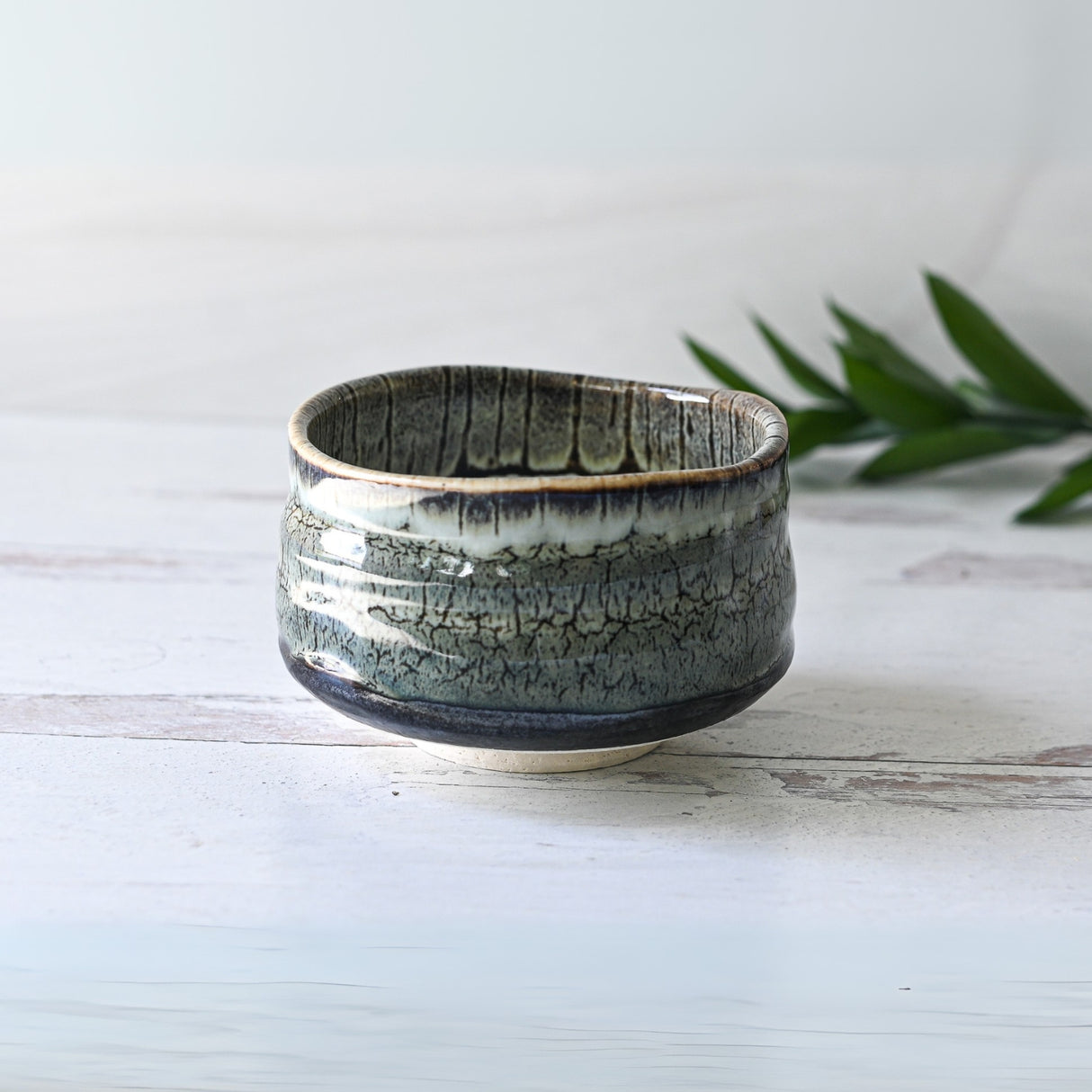The Iguchi Tenmoku Glaze Matcha Bowl, featuring a textured green and gray pattern on a white surface reminiscent of Minoyaki craftsmanship, sits beautifully next to lush green leaves.