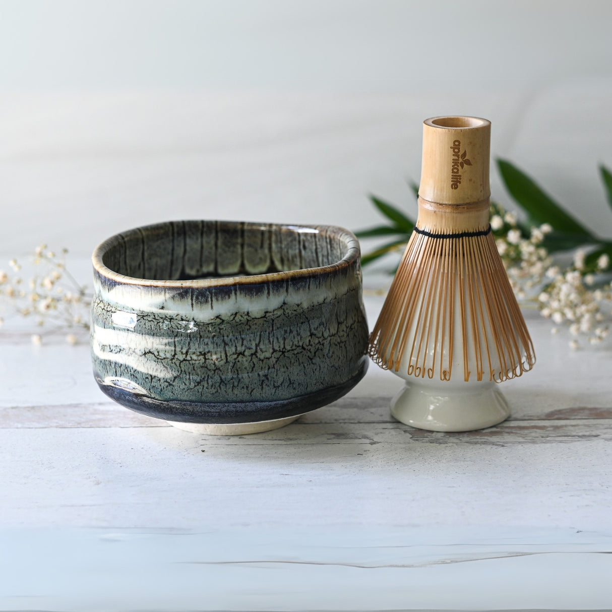 An Iguchi Tenmoku Glaze Matcha Bowl and bamboo whisk sit on a light wood surface, surrounded by greenery and small white flowers in the background.