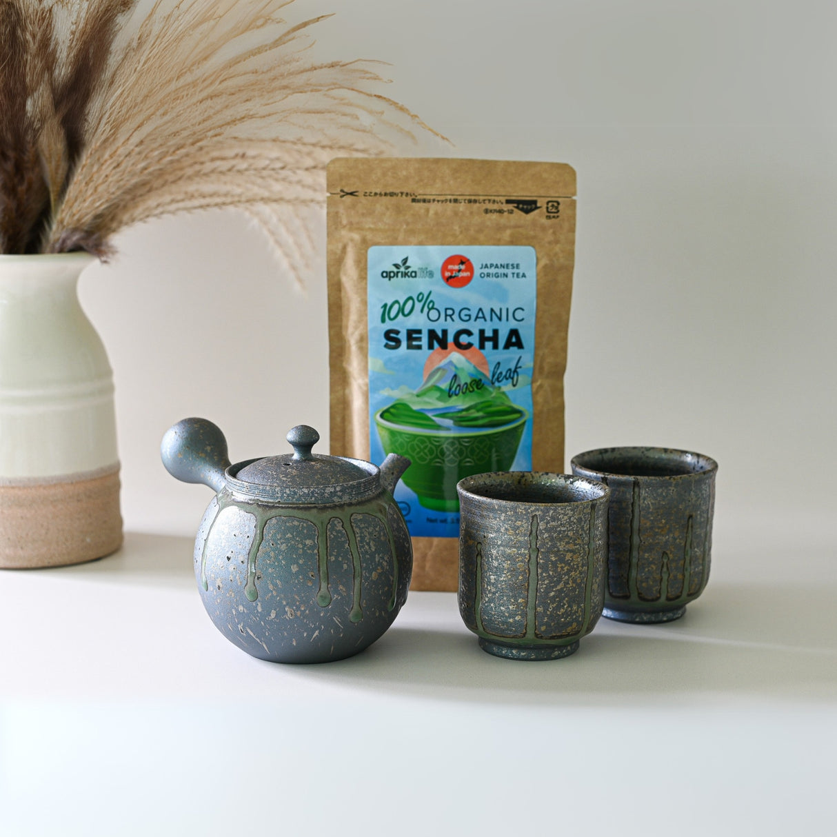 The Japanese Kyusu Set, featuring a 230ml teapot with filters and two cups, rests on a white surface alongside organic sencha tea and a pampas grass vase.