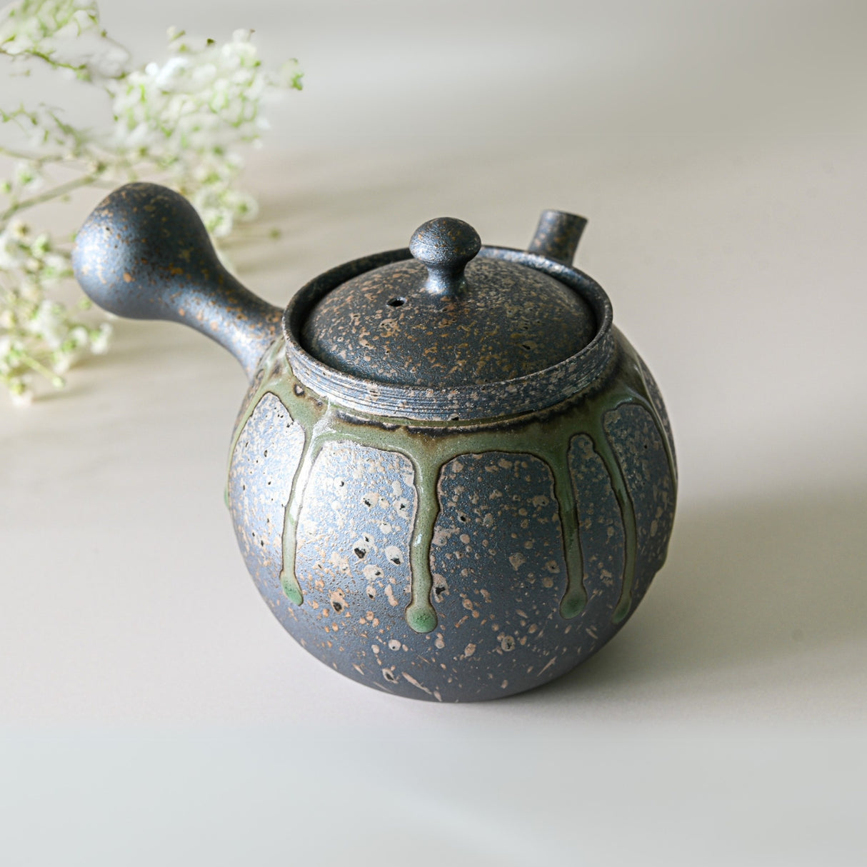 A Japanese Kyusu set, featuring a Yakishime teapot with filters (230ml) and two cups, sits beside delicate white flowers, showcasing the artistry of Tokoname ceramics.