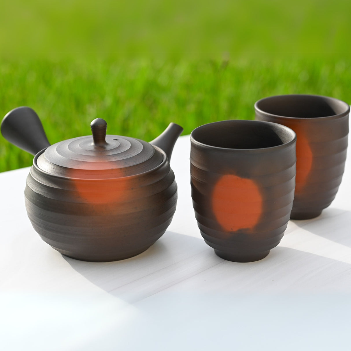The Japanese Kyusu Set - Teapot with Filters (270ml) and Two Cups sits elegantly on a white surface, its dark design with orange accents offering a striking contrast against the lush green grass backdrop.