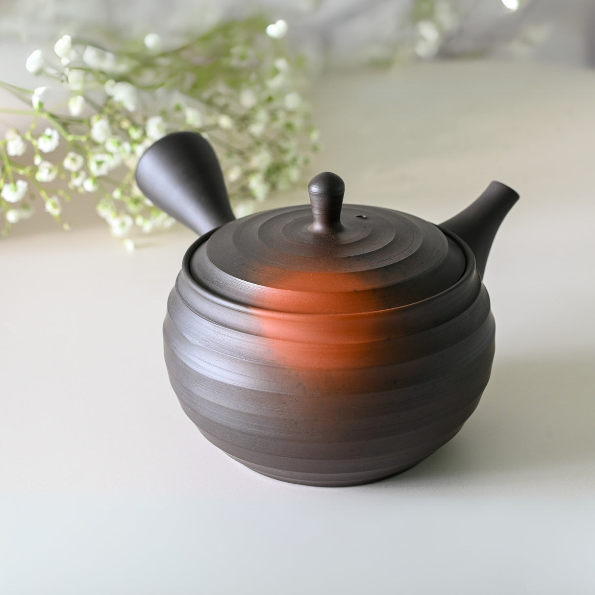 A Japanese Kyusu teapot (270ml), part of a set with filters and two cups, featuring a dark hue with a reddish tint, is elegantly surrounded by delicate white flowers on a smooth surface.