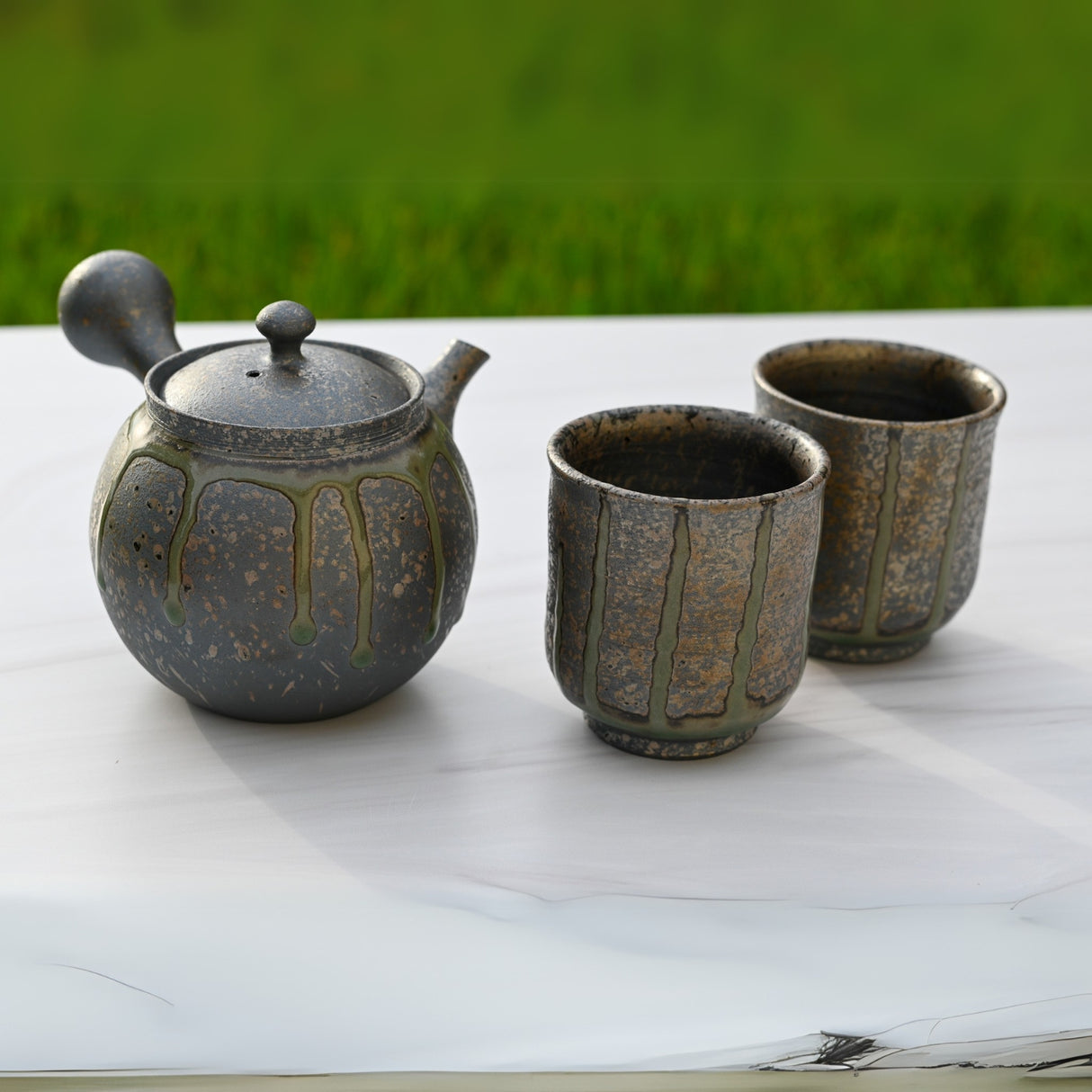 A Tokoname ceramics masterpiece: the Japanese Kyusu Set - Teapot with Filters (230ml) and Two Cups sits elegantly on a white marble table against a lush grassy backdrop.