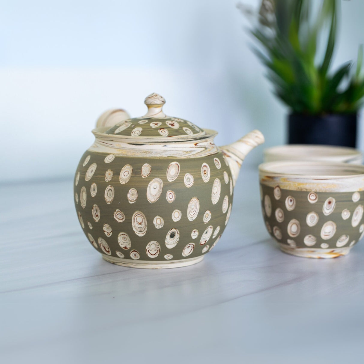 The Japanese Kyusu Set, featuring a teapot with filters (370ml) and two cups adorned with circular patterns, rests on a white surface. The set's delicate Tokoname clay hues are beautifully enhanced by a vibrant plant in the background, inviting tranquility over green tea.