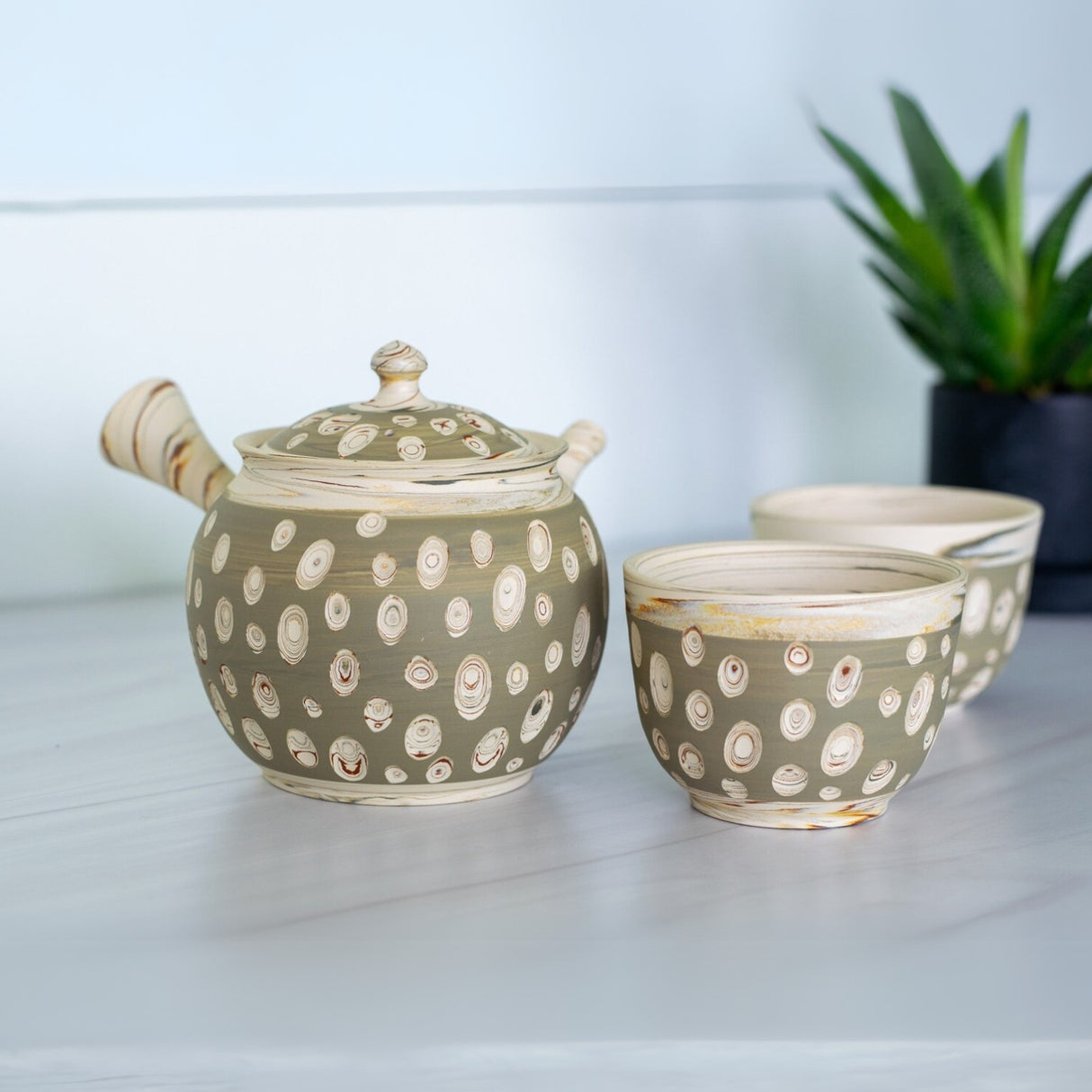 A Japanese Kyusu Set, featuring a 370ml teapot with filters and two ceramic cups, sits elegantly on the table, complemented by a small potted plant—perfect for savoring green tea.