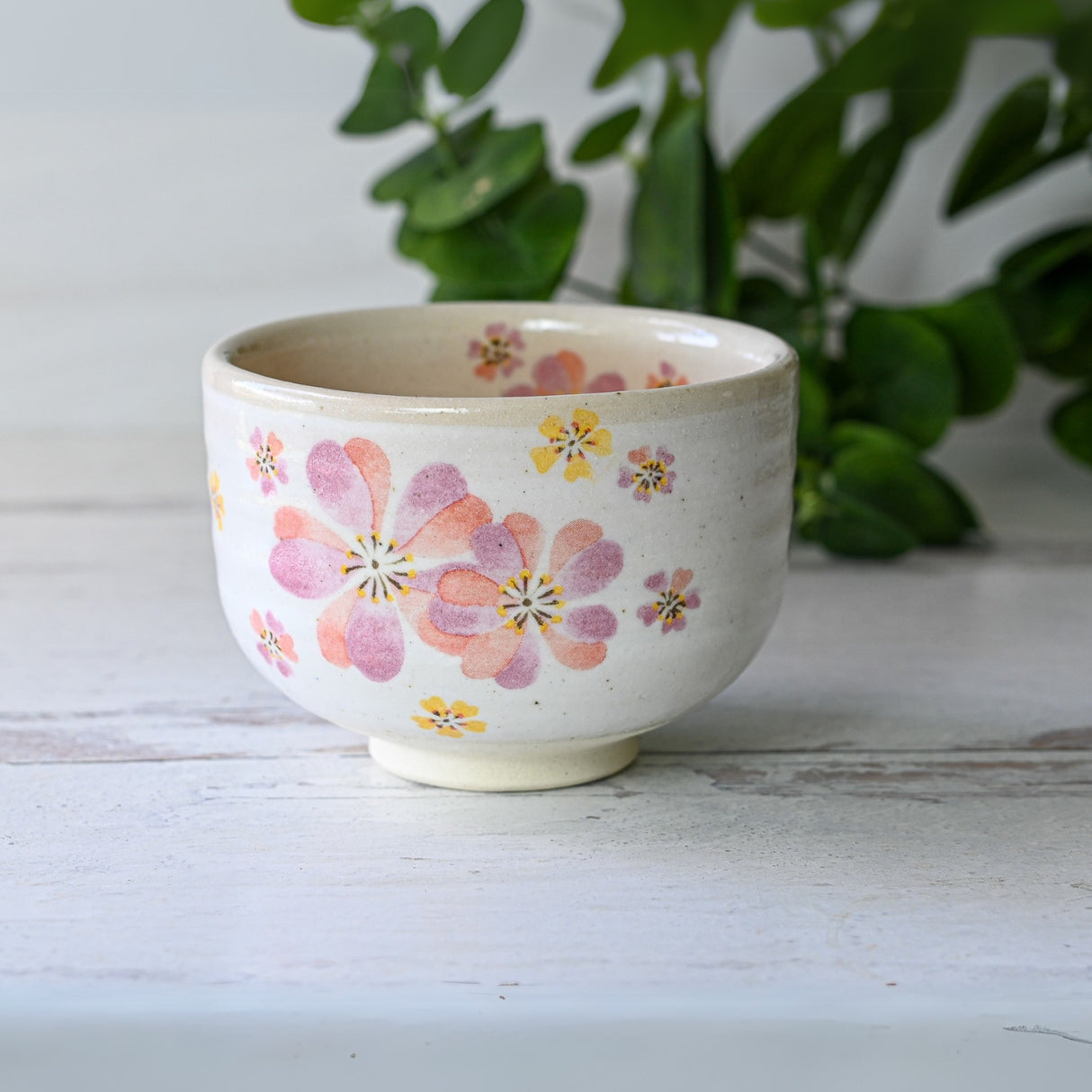 The Japanese Pink Sakura Ceramic Cup, reminiscent of Minoyaki craftsmanship, features pink flower designs on white. It sets a serene scene with greenery in the background.
