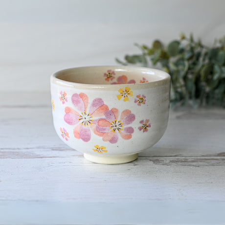 The Japanese Pink Sakura Ceramic Matcha Bowl with a floral design rests elegantly on a wooden surface, surrounded by lush greenery.