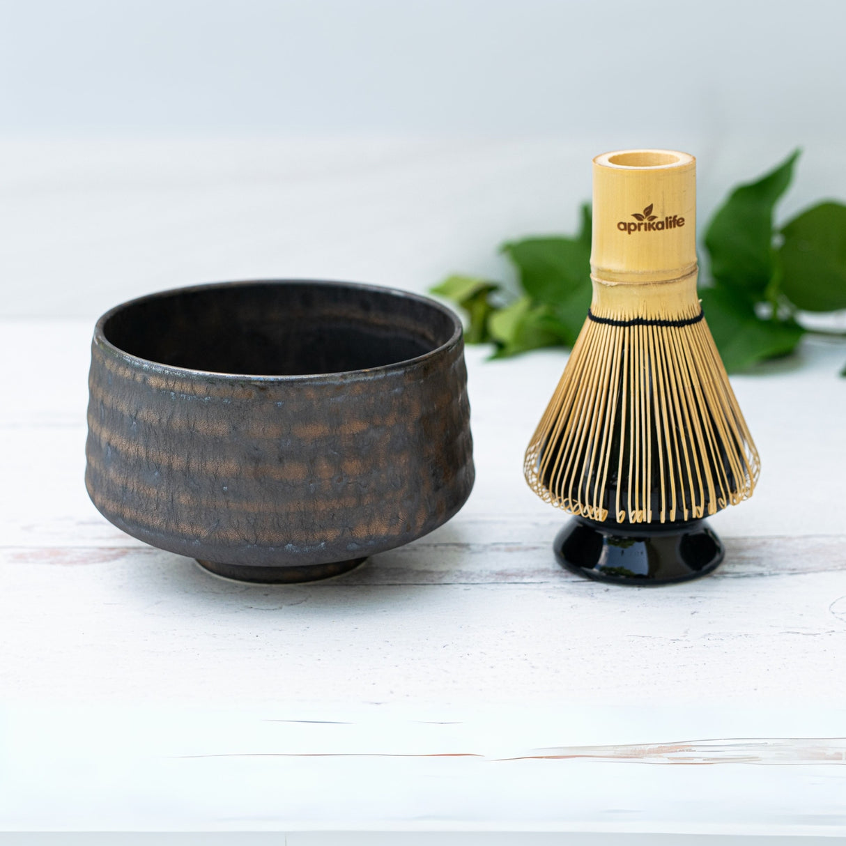 The Kaneni Gold Crystal Ceramic Matcha Bowl and a bamboo whisk are set on a white wooden surface, with green leaves in the background.