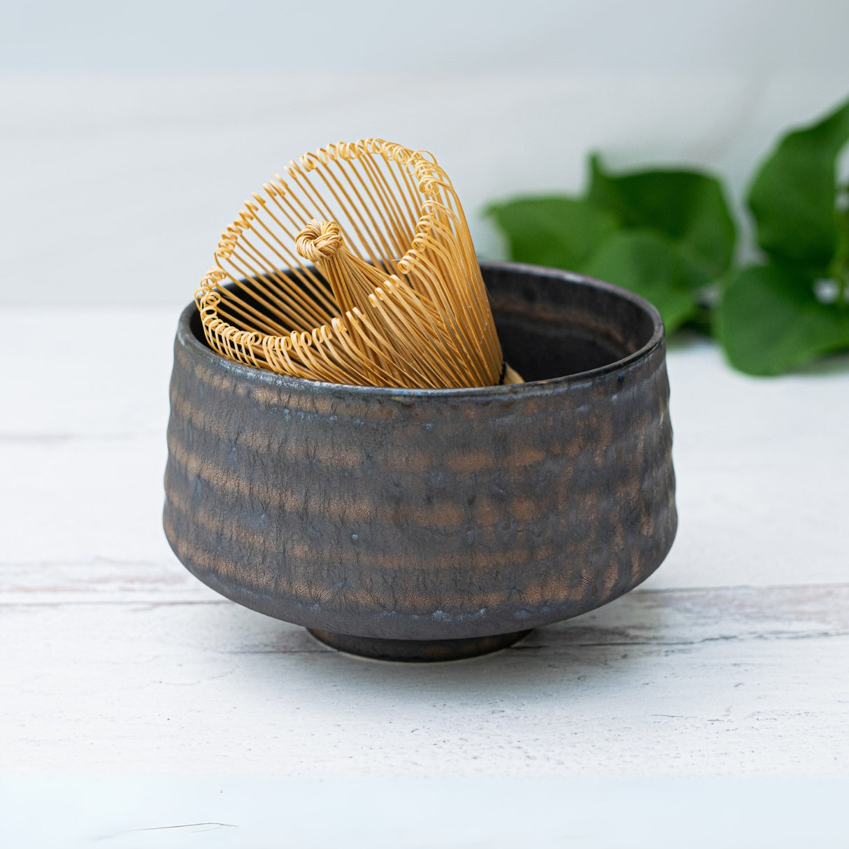 The Kaneni Gold Crystal Ceramic Matcha Bowl, with a bamboo matcha whisk and holder, showcases Minoyaki craftsmanship—set against greenery, it’s an elegant addition to your matcha accessory collection.