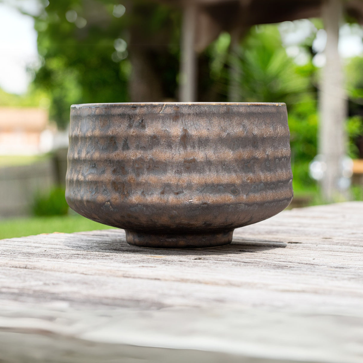 The Kaneni Gold Crystal Ceramic Matcha Bowl, featuring rustic Minoyaki pottery techniques, sits on a wooden table, with soft greenery framing the scene.