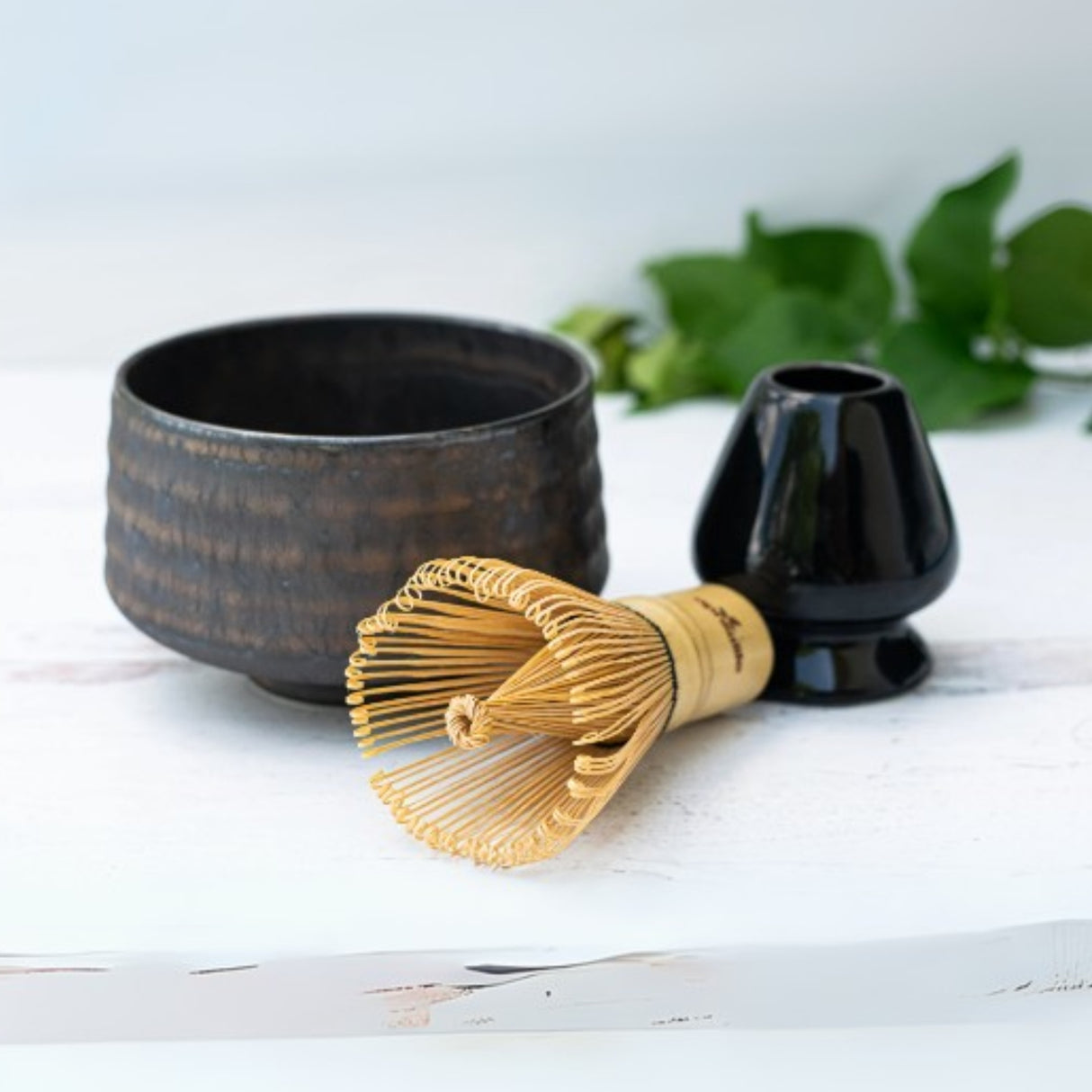The Kaneni Gold Crystal Ceramic Matcha Bowl is crafted using Minoyaki pottery techniques and includes a bamboo whisk, black bowl, and holder on a white surface with green leaves in the background.