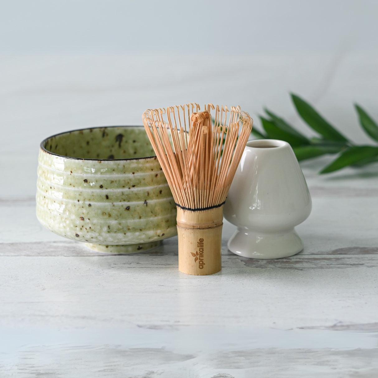 The Kaneni White Ceramic Matcha Bowl, Bamboo Matcha Whisk, Whisk Holder, and Organic Japanese Matcha Set is displayed on a light wooden surface with green leaves. The bowl is crafted using Minoyaki techniques.