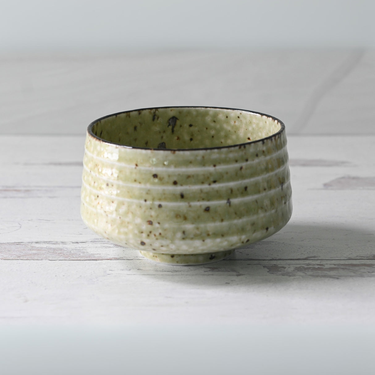 The Kaneni White Ceramic Matcha Bowl, with brown speckles and textured light green glaze, crafted via Minoyaki techniques, sits on a light wooden surface alongside its bamboo whisk, whisk holder, and organic Japanese matcha.