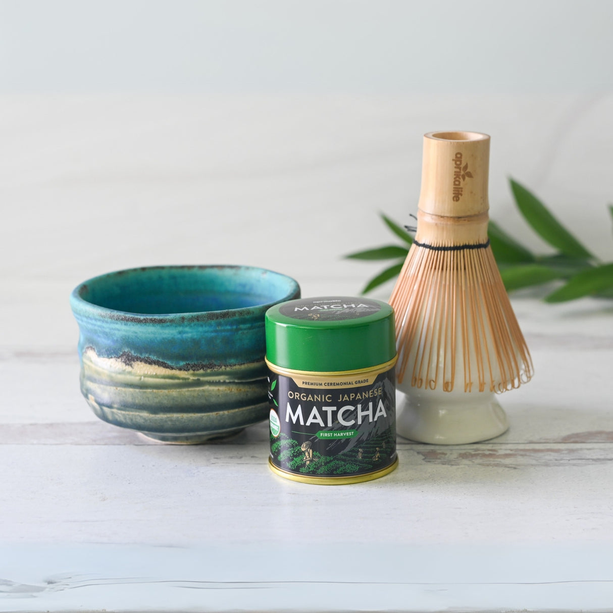 A Marusho Blue Matcha Bowl set, including a bamboo matcha whisk and holder, is placed on a light surface with lush greenery in the background.