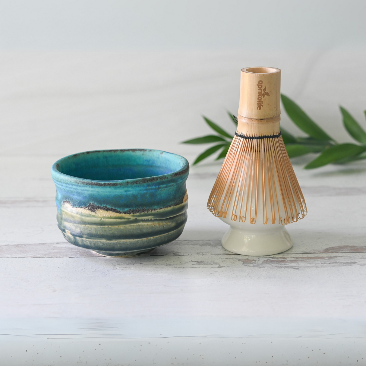 A Marusho Blue Matcha Bowl and Bamboo Matcha Whisk from the Organic Japanese Matcha Set rest elegantly on a white wood surface, framed by green leaves.