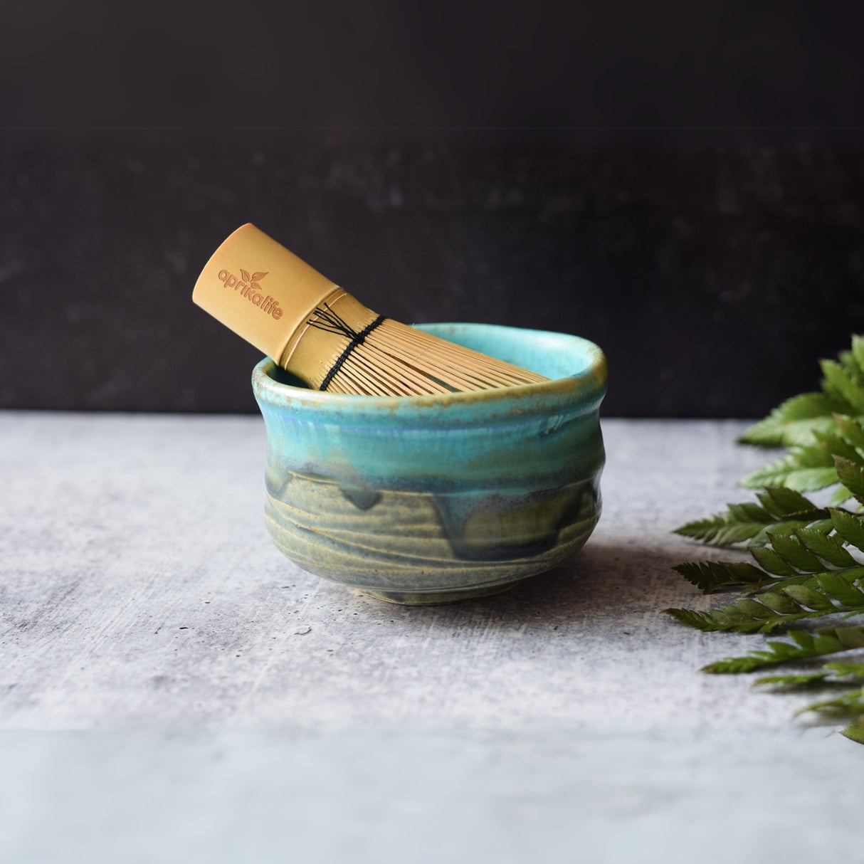 Marusho Blue Matcha Bowl with a bamboo whisk rests on a textured surface next to green leaves.