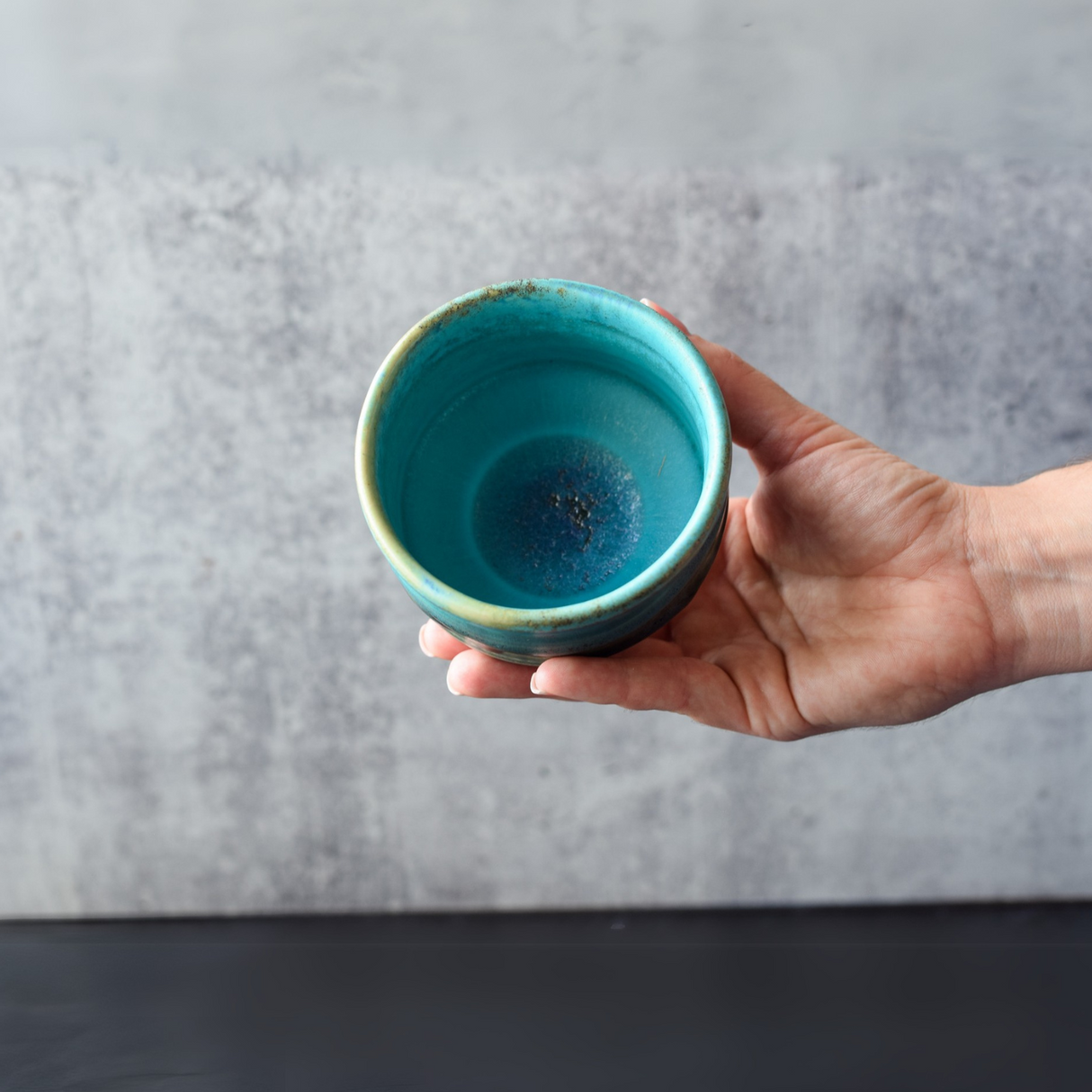 A hand holds a small Marusho Blue Matcha Bowl with a textured surface against a gray background, reminiscent of traditional Japanese pottery.