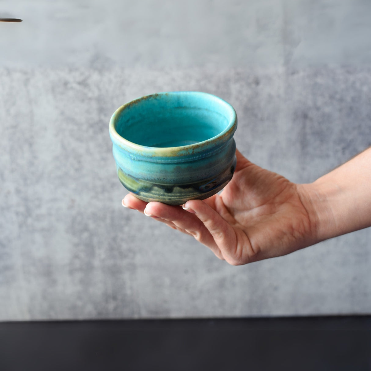 A hand holds a Marusho Blue Matcha Bowl against a gray, textured background.