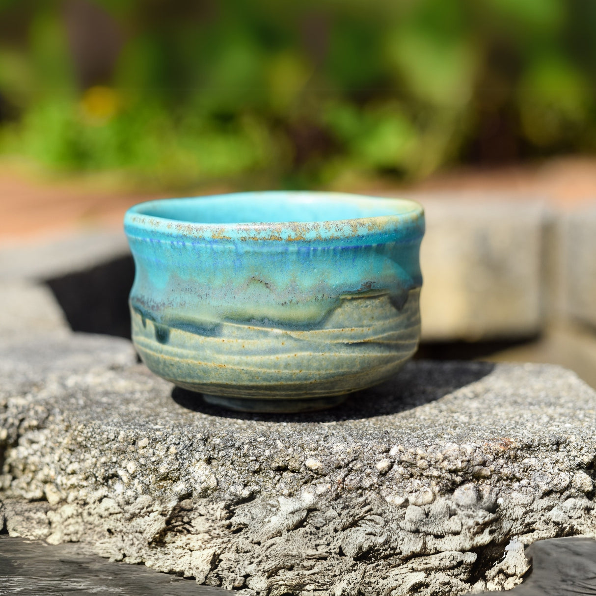 The Marusho Blue Matcha Bowl from the Matcha Set, featuring rustic earthy tones, rests on a textured stone surface outdoors, showcasing the artistry of Minoyaki Japanese pottery.