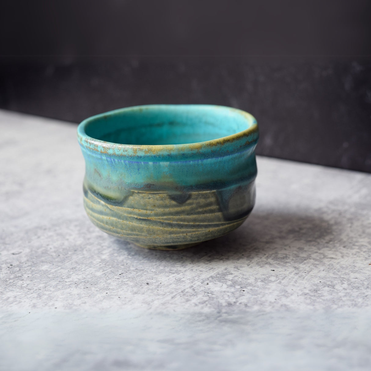 A Marusho Blue Matcha Bowl from the Bamboo Matcha Whisk Set, featuring turquoise and earthy glaze, rests on a textured surface, capturing the essence of traditional Japanese pottery.