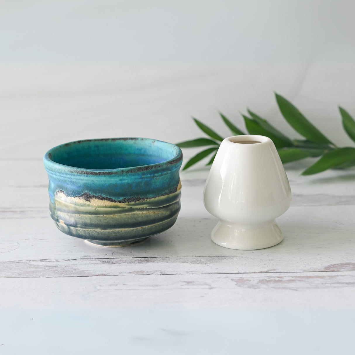 A Marusho Blue Matcha Bowl and a white ceramic vase rest gracefully on a light wooden surface, surrounded by green leaves.