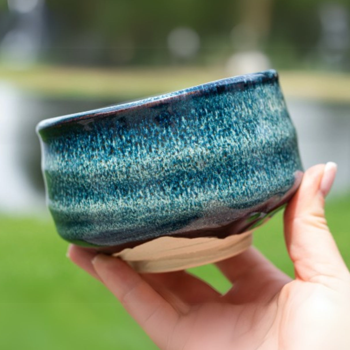 A hand holds the Maruyoshi Navy Sea Ceramic Matcha Bowl outdoors against a backdrop of blurred greenery.