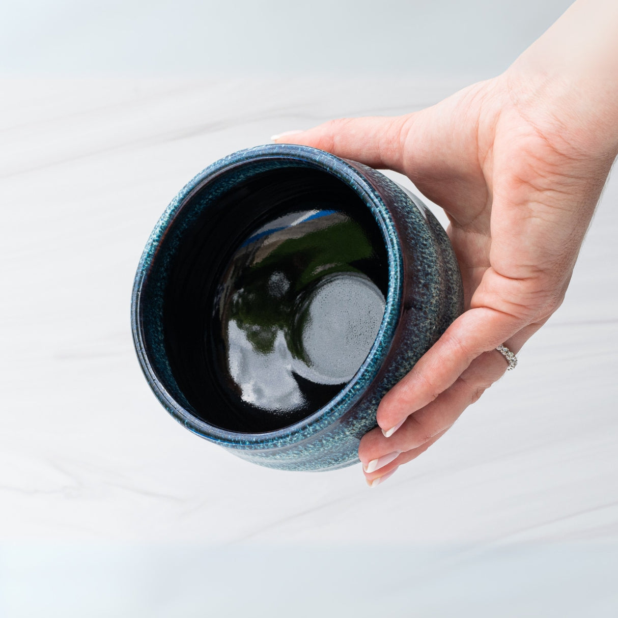 A hand cradles the Maruyoshi Navy Sea Ceramic Matcha Bowl, with a glossy black interior contrasting against a light setting, evoking tranquility and the artful ritual of matcha tea.