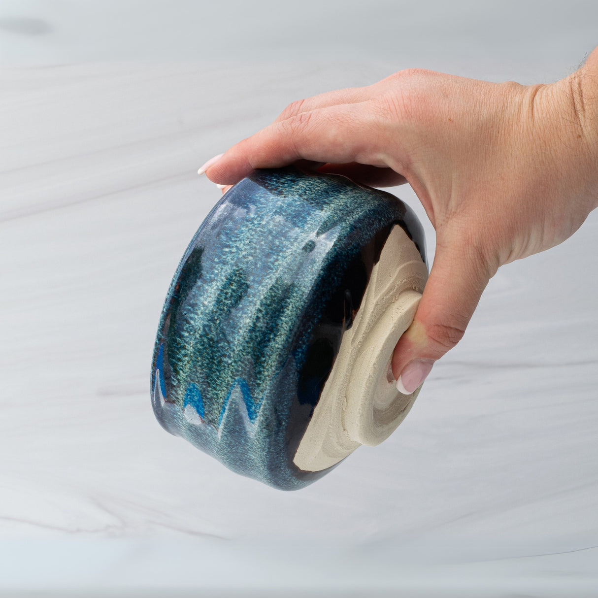 A hand gently cradles the Maruyoshi Navy Sea Ceramic Matcha Bowl, showcasing its blue-green hue and textured design with an unglazed base.